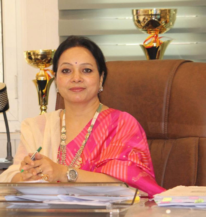 Woman in pink sari sits at desk signing papers with a pen; trophies on the shelf behind her.