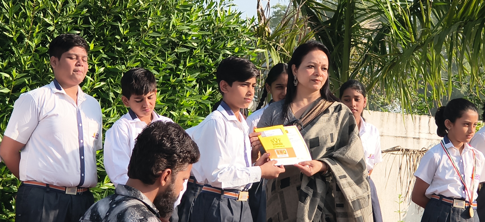Teacher hands a certificate to a student during an outdoor awards ceremony with classmates in uniform nearby.