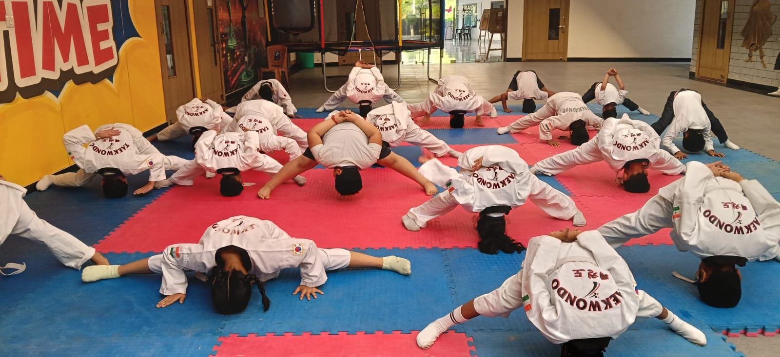 Group of children in white taekwondo uniforms doing backbend stretches on blue and pink foam mats in a gym.